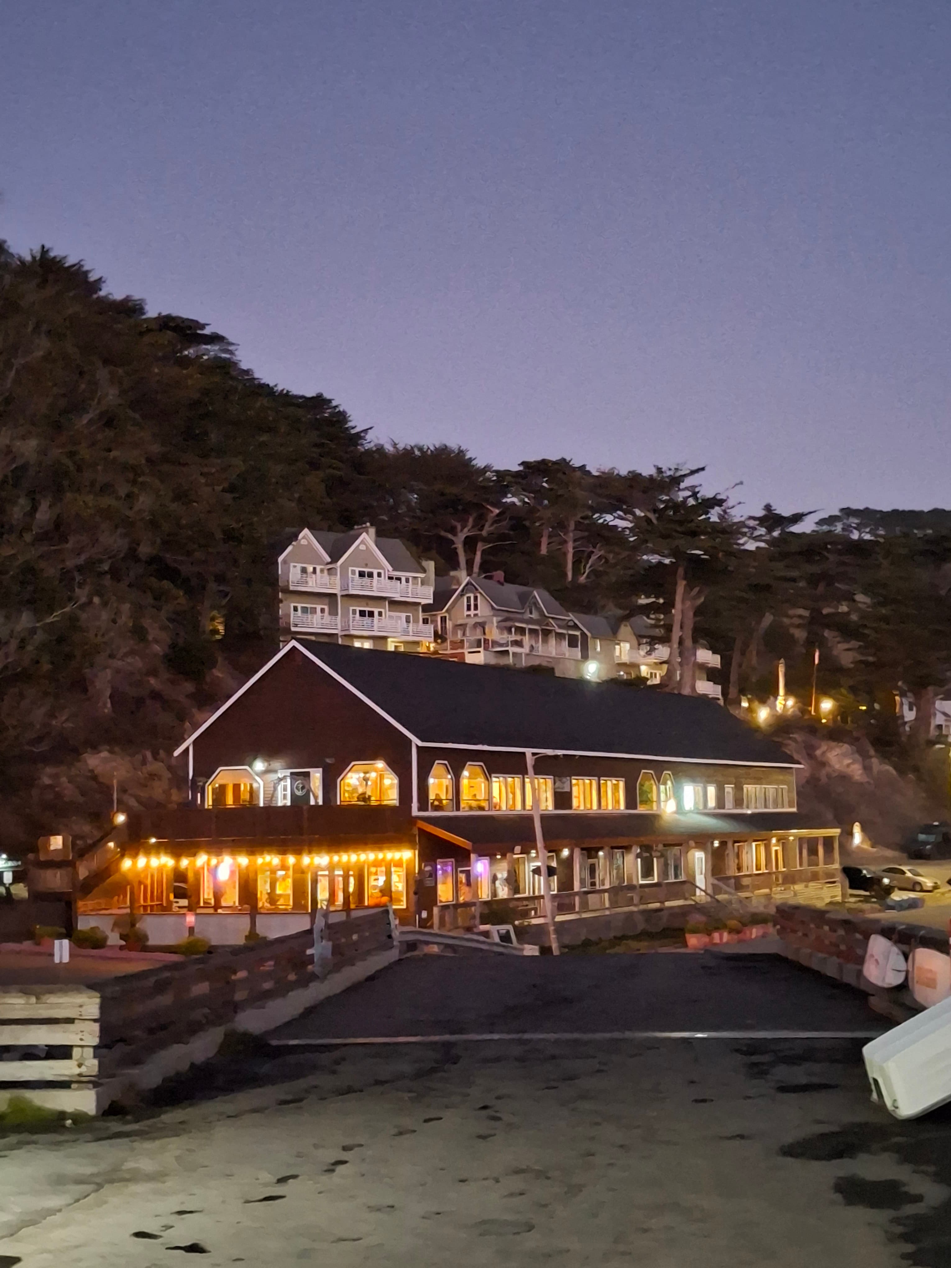 A coastal restaurant and hillside homes glowing at dusk, seen from the beach below