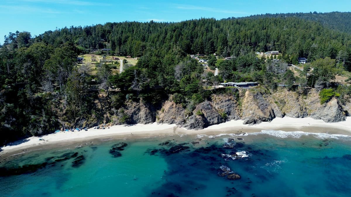 Aerial view of homes on a forested bluff above a crescent white sand beach with turquoise, rock-lined water.