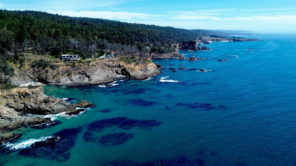 Aerial view of a single home on a forested rocky bluff overlooking deep blue open ocean and a rugged coastline.