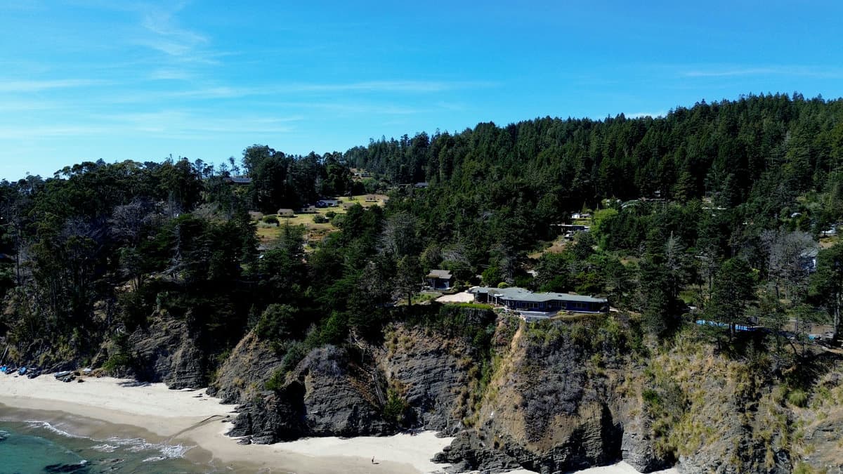 Aerial close-up of a modern home on a dark rocky cliff above a white sand beach, surrounded by forest.