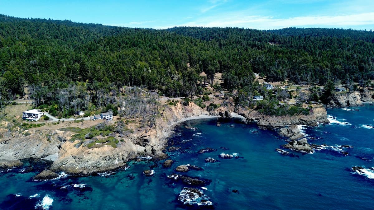 Aerial view of two homes on a grassy clifftop overlooking a rock-enclosed turquoise cove.
