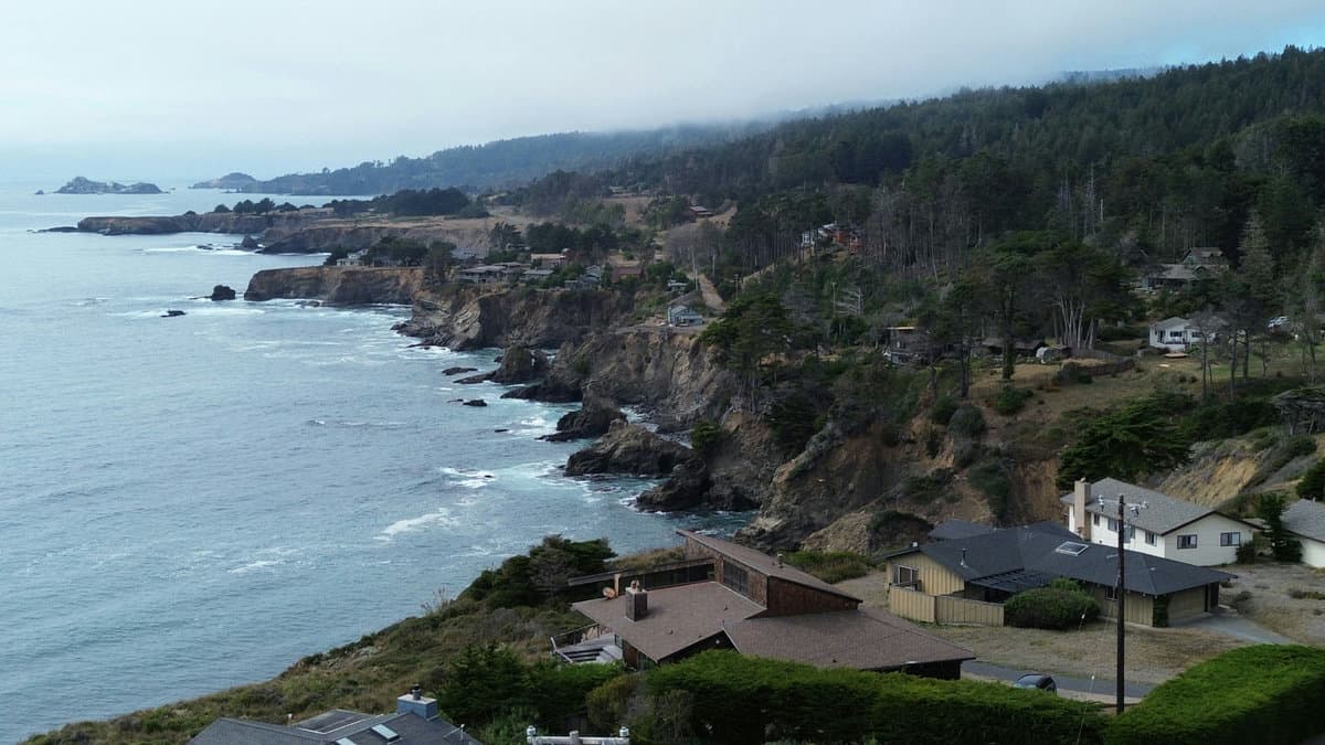 Aerial view of coastal neighborhood rooftops and a rugged shoreline receding into evening fog.